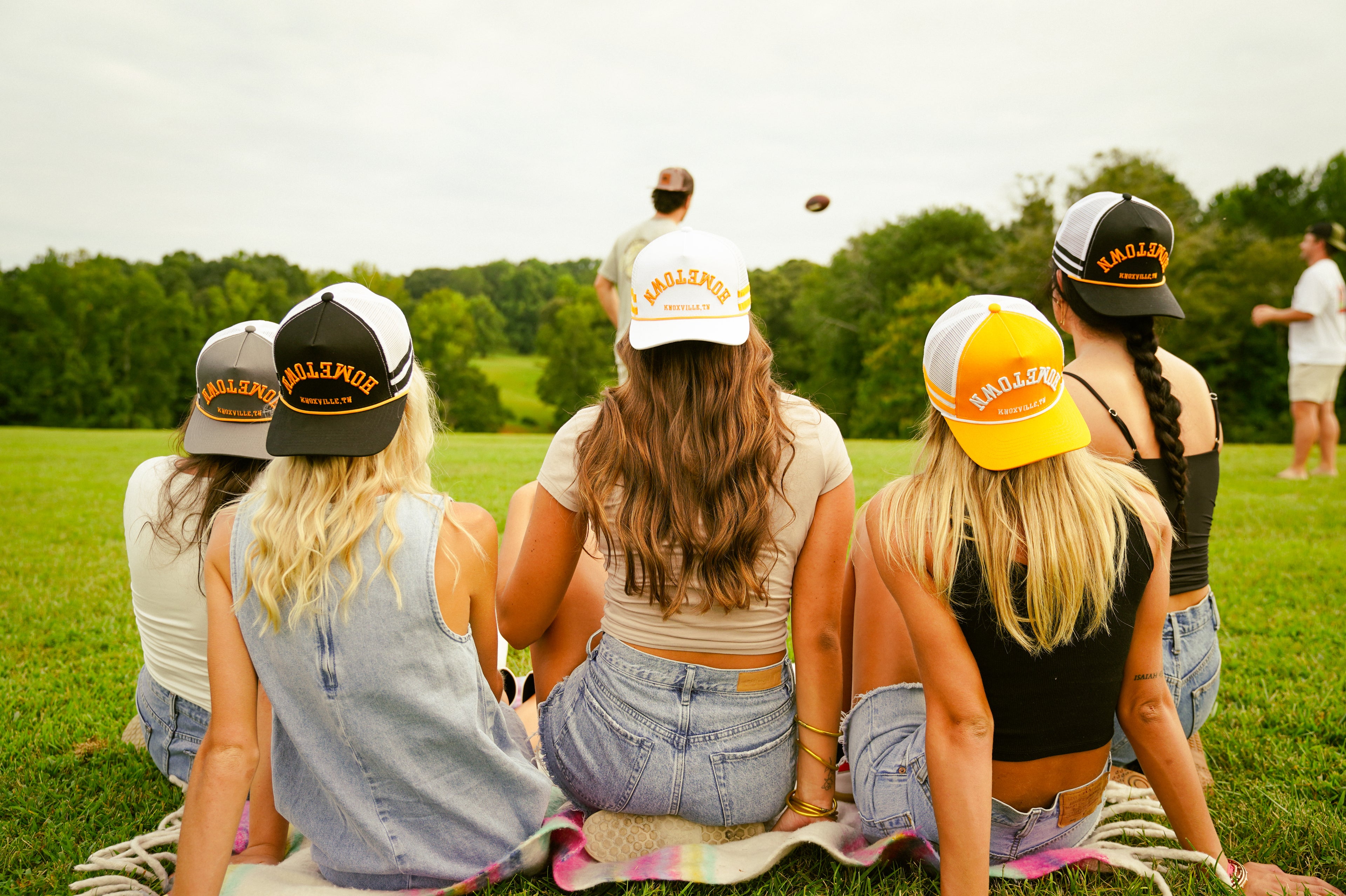 Group of people wearing Hometown baseball caps sitting on the grass with trees in the background