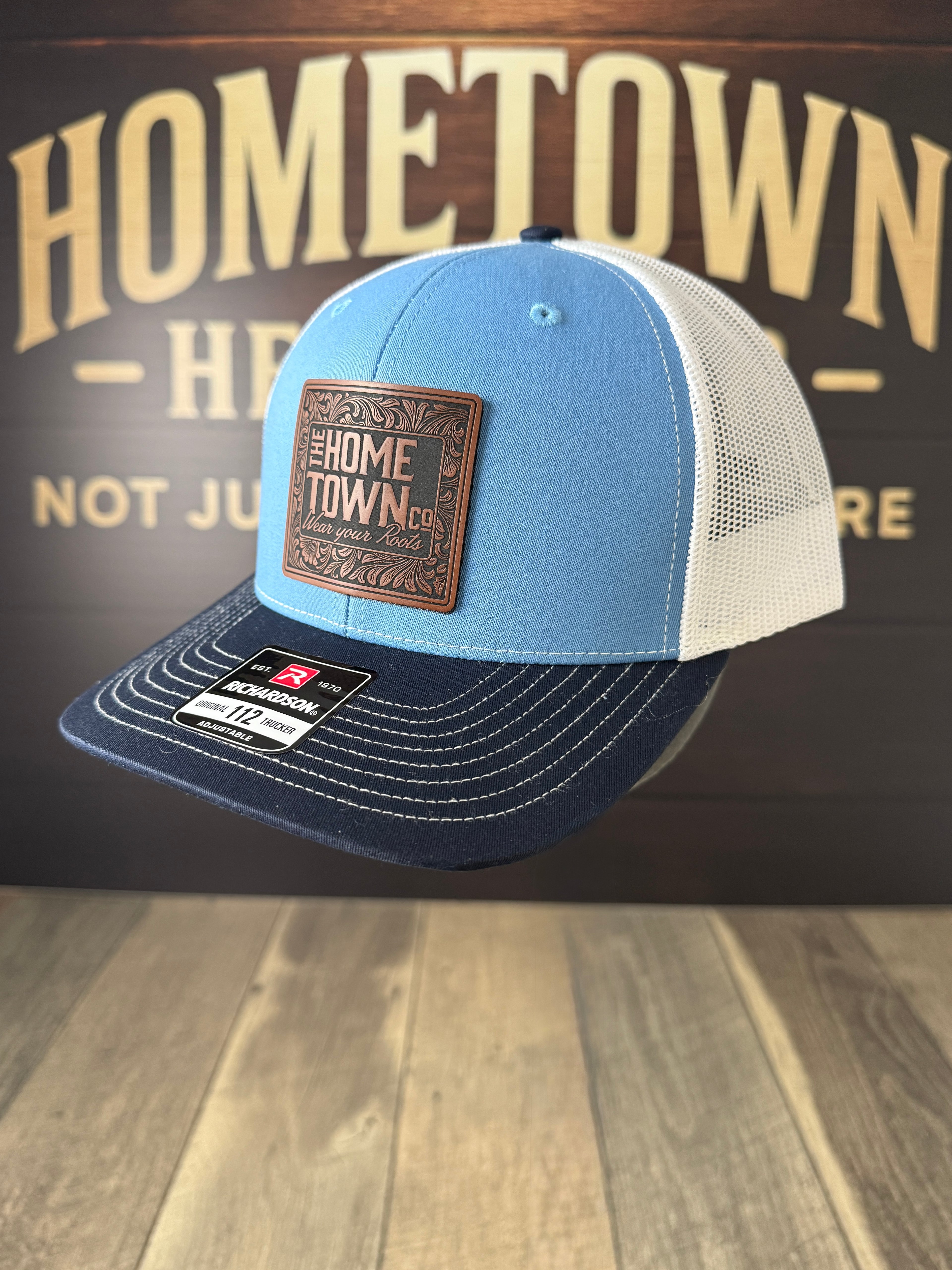 Blue and navy trucker hat with leather patch on a wooden surface with 'Hometown' background