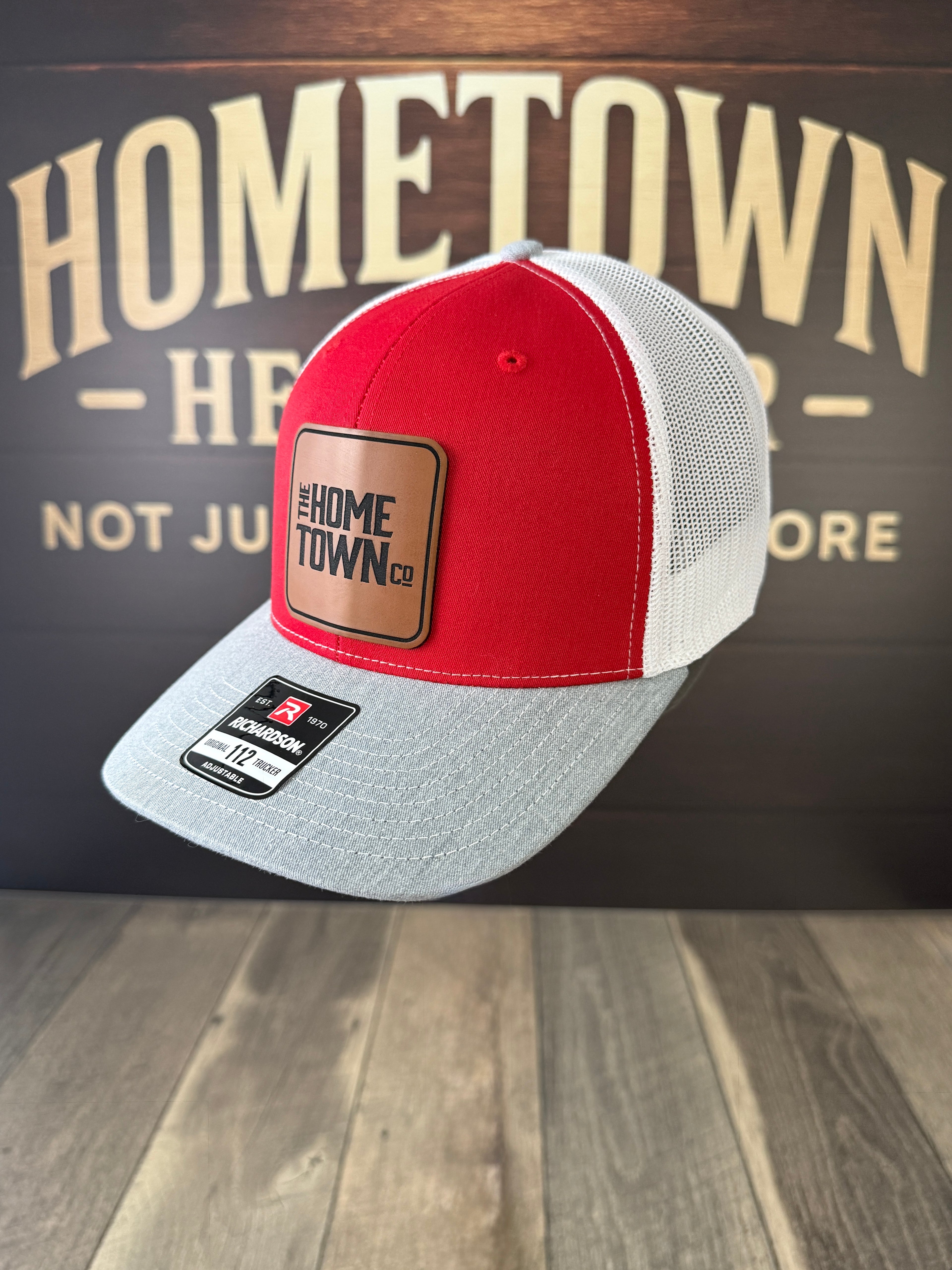 Red and gray cap with leather patch on a wooden surface with 'Hometown' background