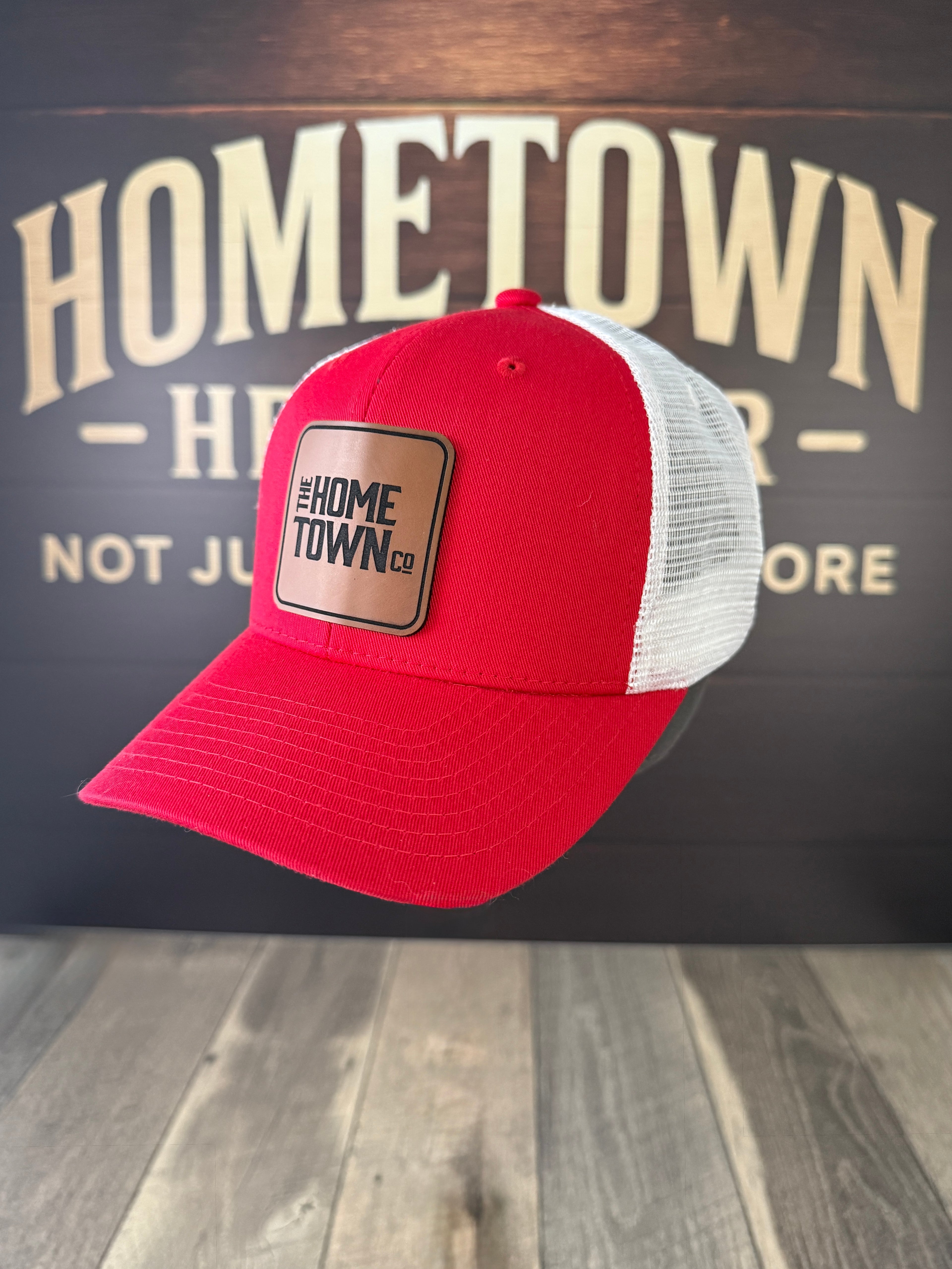 Red and white trucker hat with a patch on a wooden surface with 'Hometown' branding in the background.