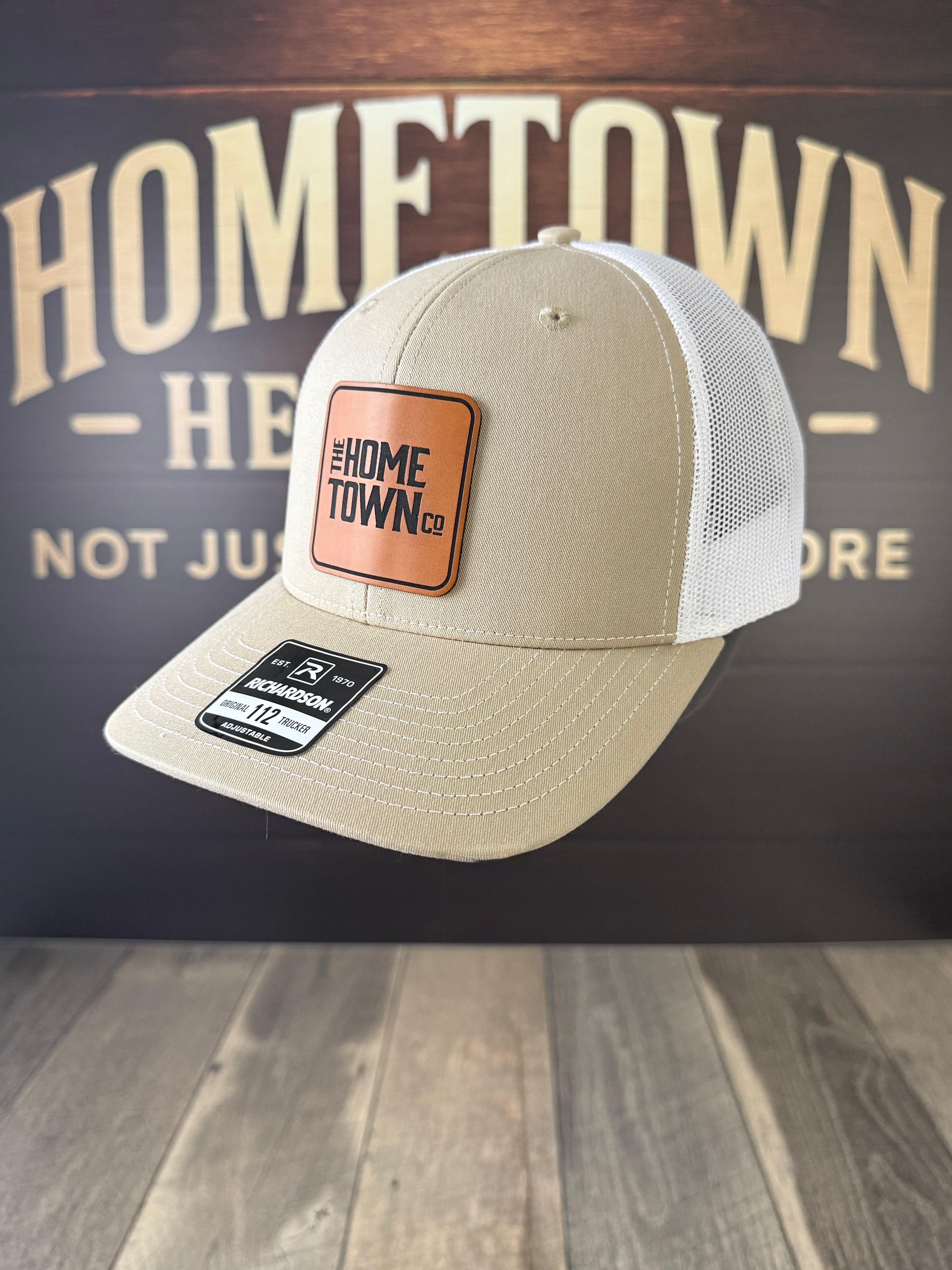 Beige cap with a brown patch on a wooden surface with 'Hometown' branding in the background.